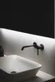 Waschbecken mit einer schwarzen Wandarmatur von Ideal Standard. Die Armatur hat einen langen Auslauf und ist direkt an der Wand montiert. Helle Wand mit sanfter Beleuchtung im Hintergrund.